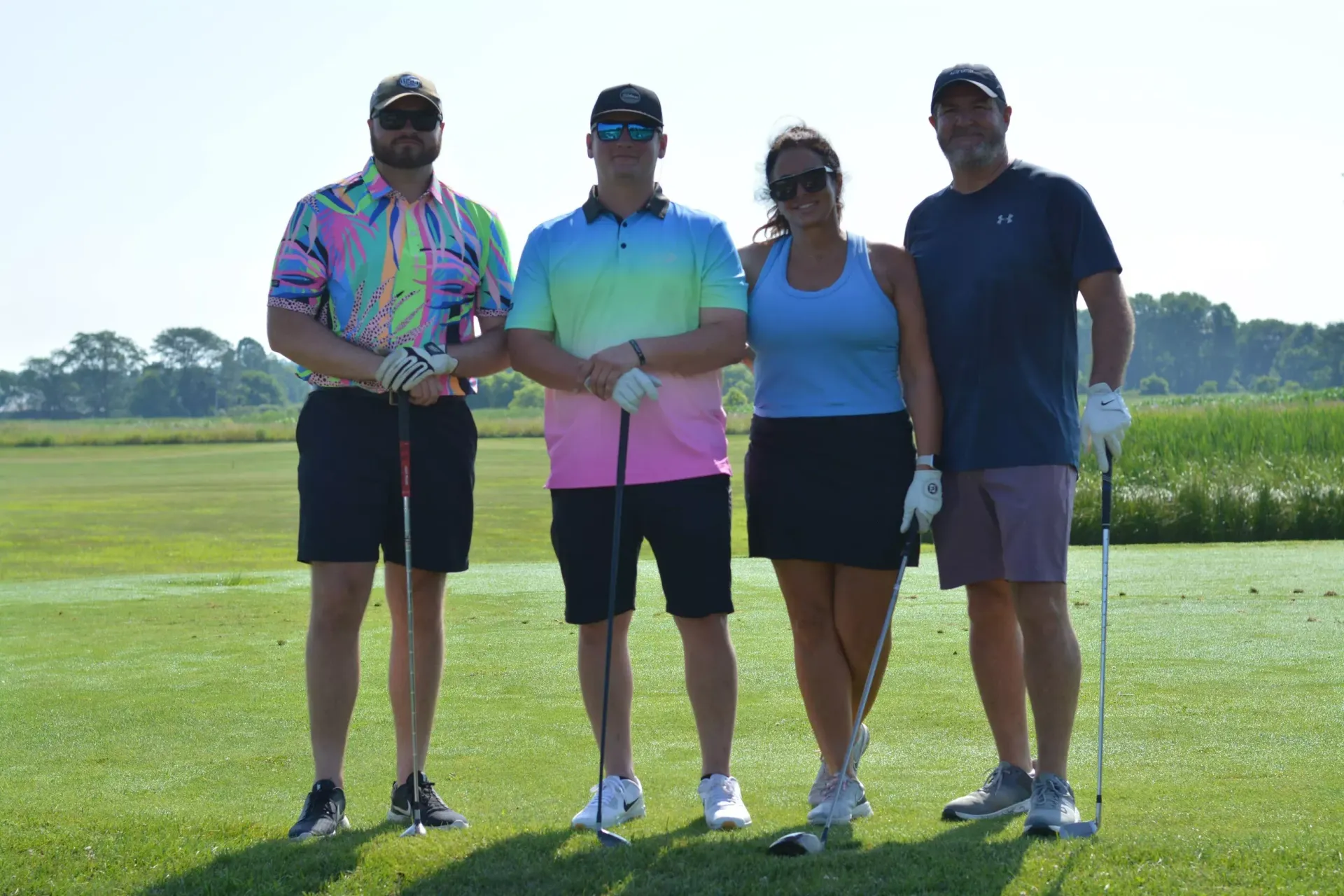 Four golfers standing on a sunny golf course with clubs and a trophy.