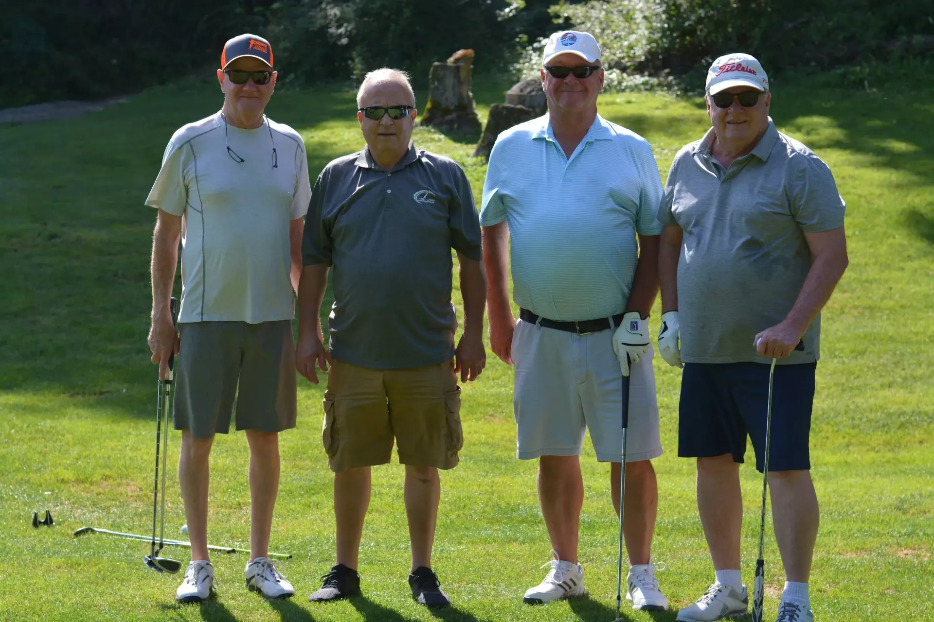 Four golfers standing together on a sunny golf course, holding clubs and wearing caps.