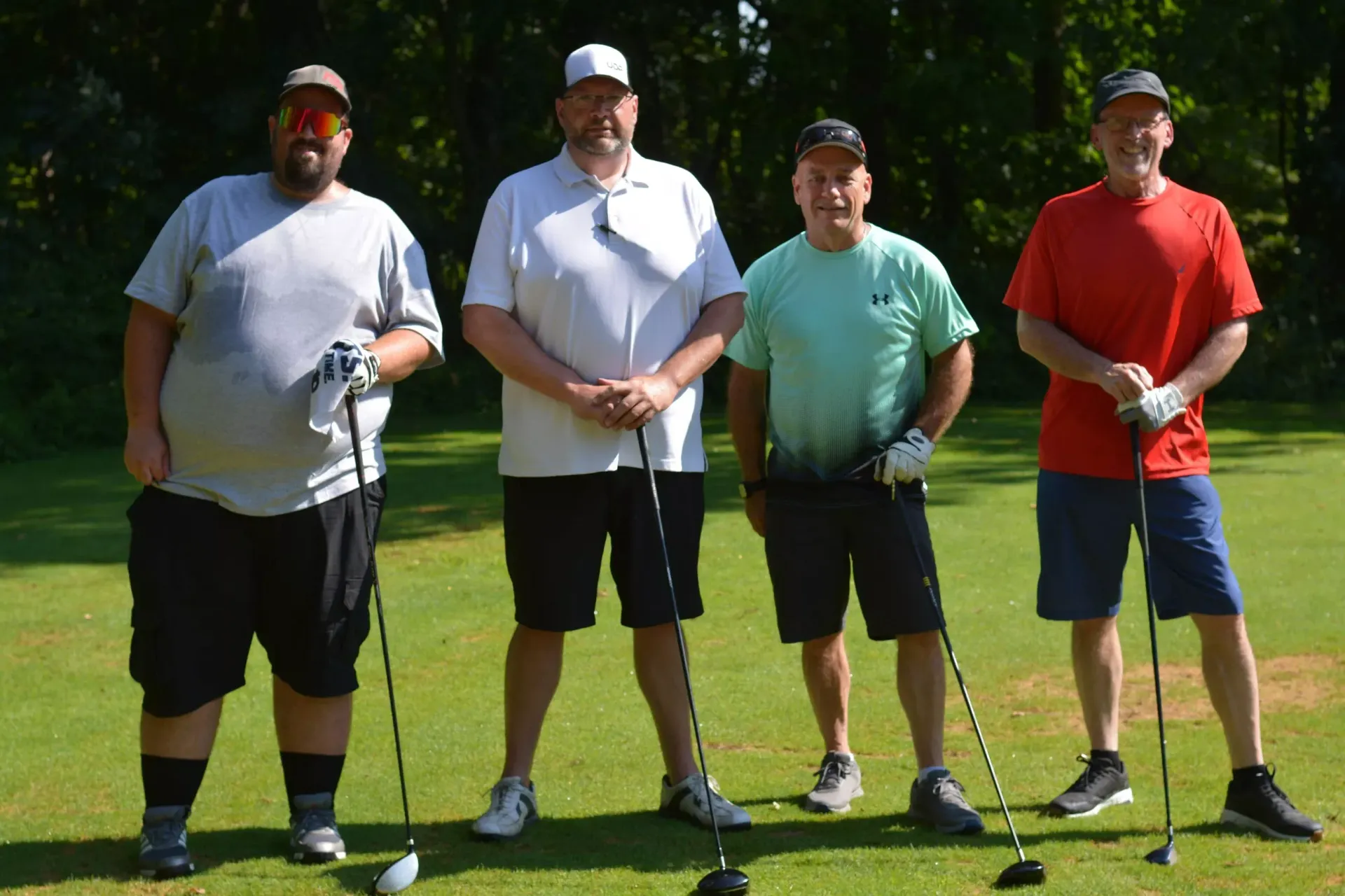 Four golfers standing on a green fairway with clubs, wearing casual golf attire and caps.