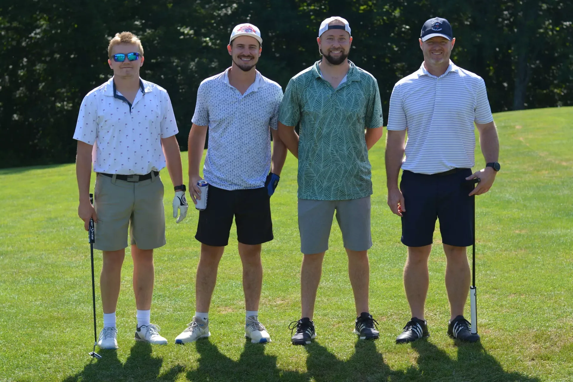 Four men standing on a sunny golf course, wearing polos and shorts, posing side by side.