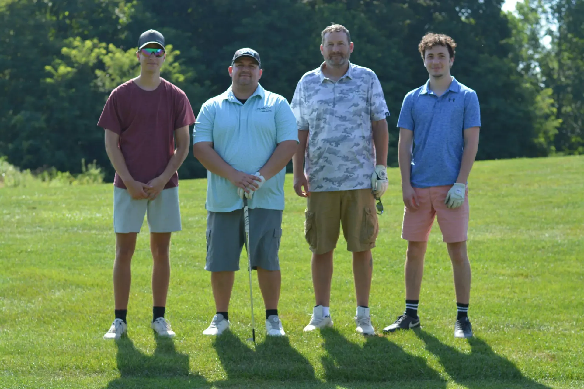 Four men standing on a sunny golf course, wearing casual shirts, shorts, and caps.