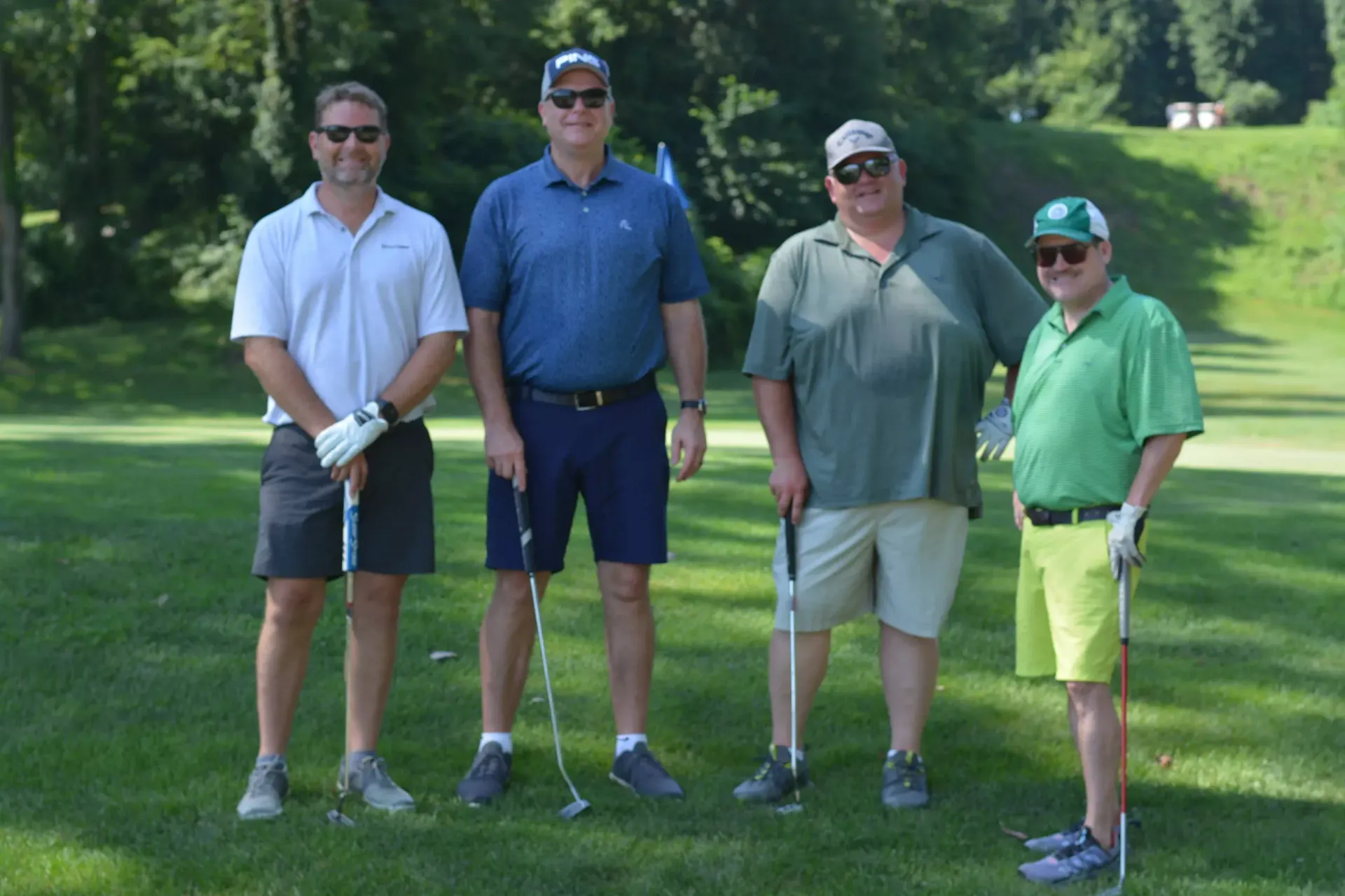 Four golfers standing on a grassy course, holding clubs, with trees in the background.