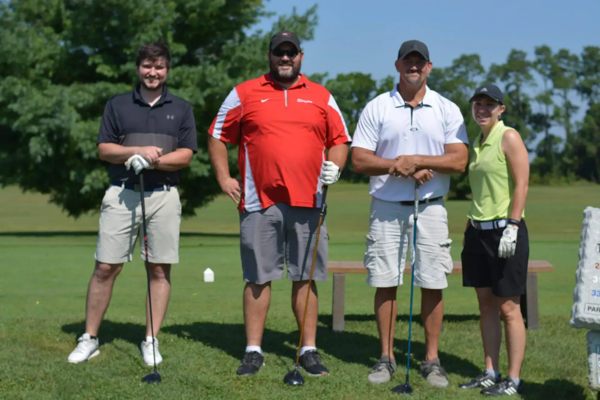 Four golfers posing on a tee box, holding clubs, with a green fairway in the background.
