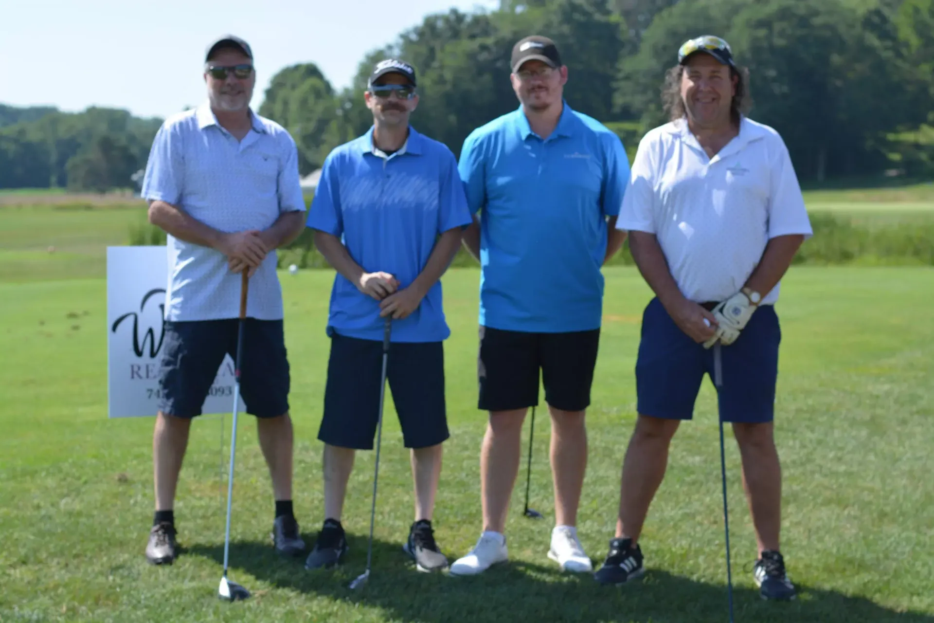 Four men stand on a golf course holding clubs, posing for a photo near a tee sign.