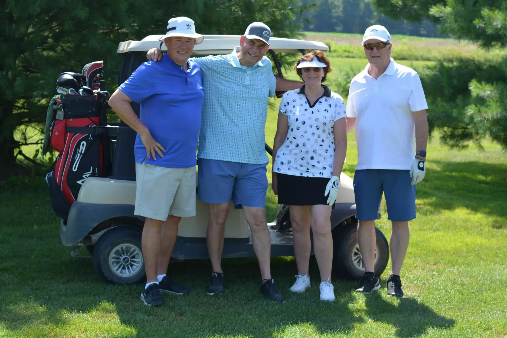 Four people posing on a golf course beside a golf cart and bags, smiling in sunlight.