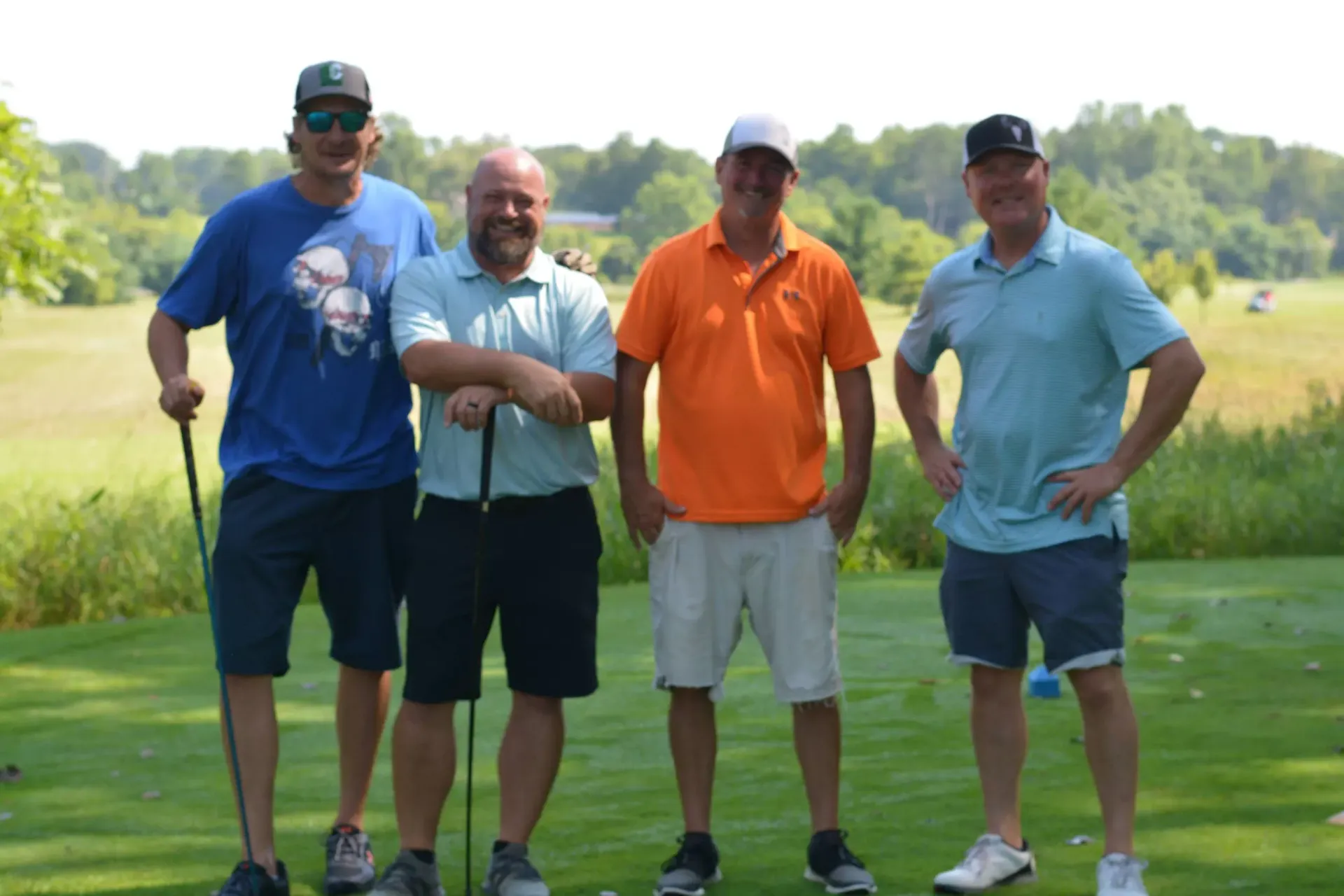 Four men standing on a golf course, holding clubs and posing outdoors.