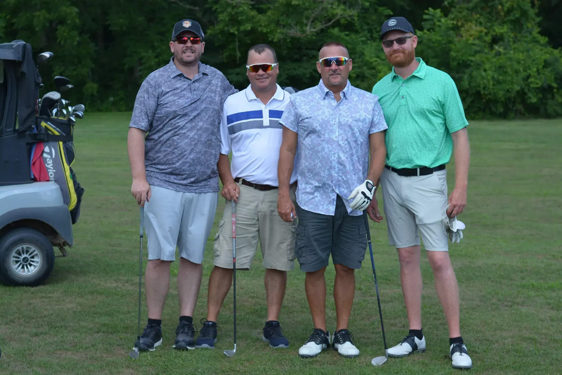 Four golfers posing on a grassy course, with a golf cart partly visible at left.