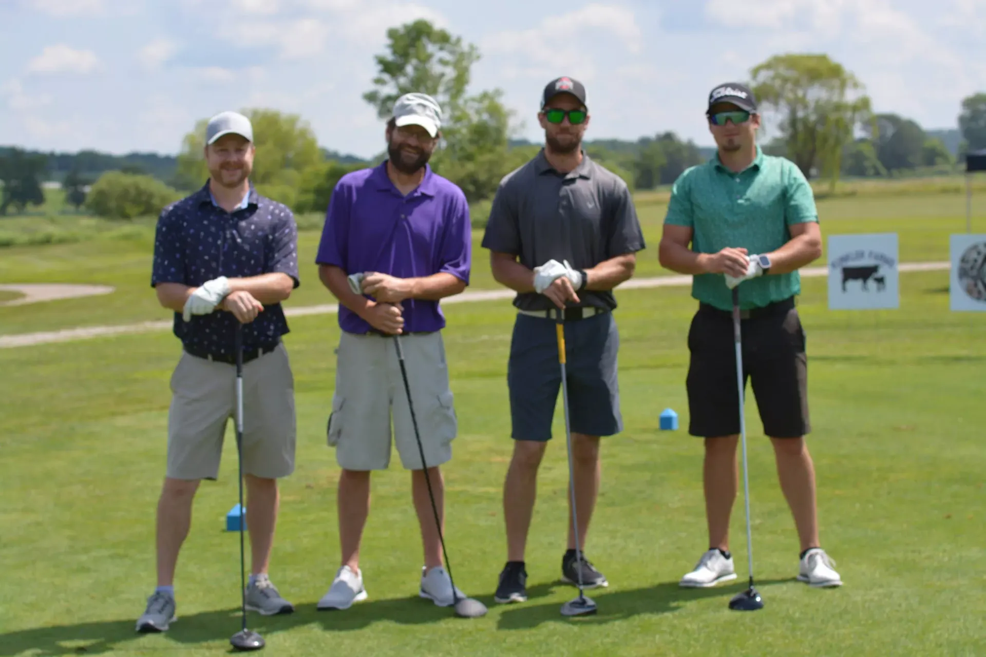 Four golfers posing on a green course with clubs, standing side by side.
