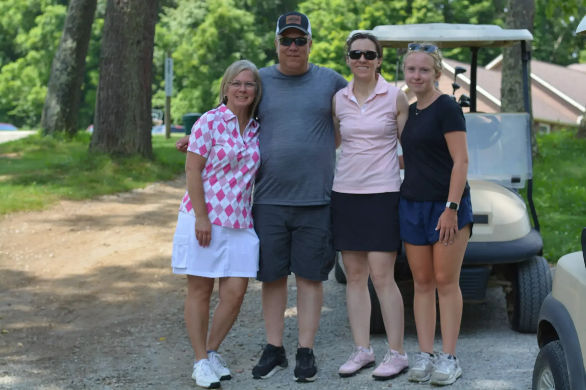 Four people stand smiling beside a golf cart on a wooded path.