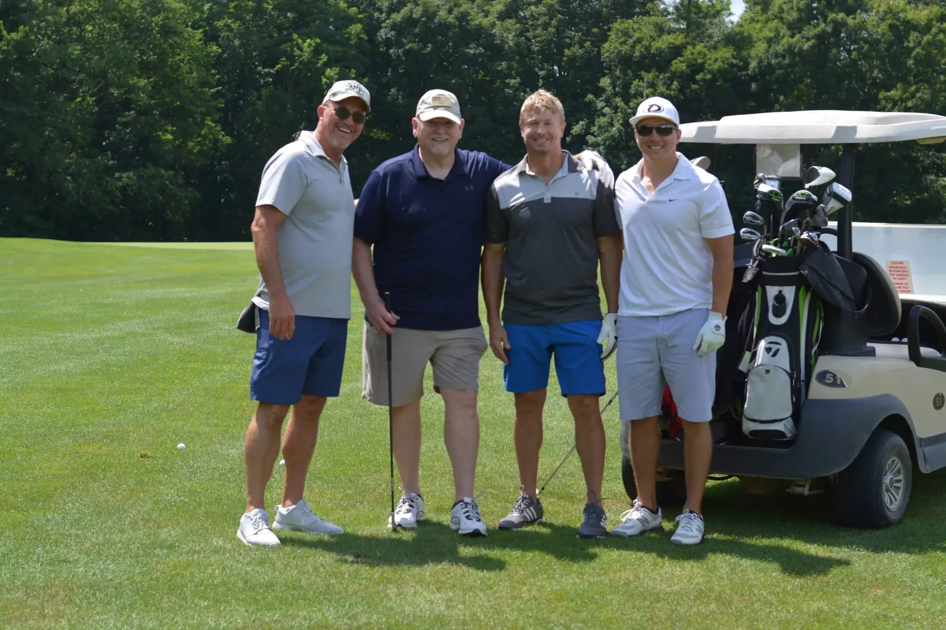 Four people standing on a golf course beside a golf cart on a sunny day