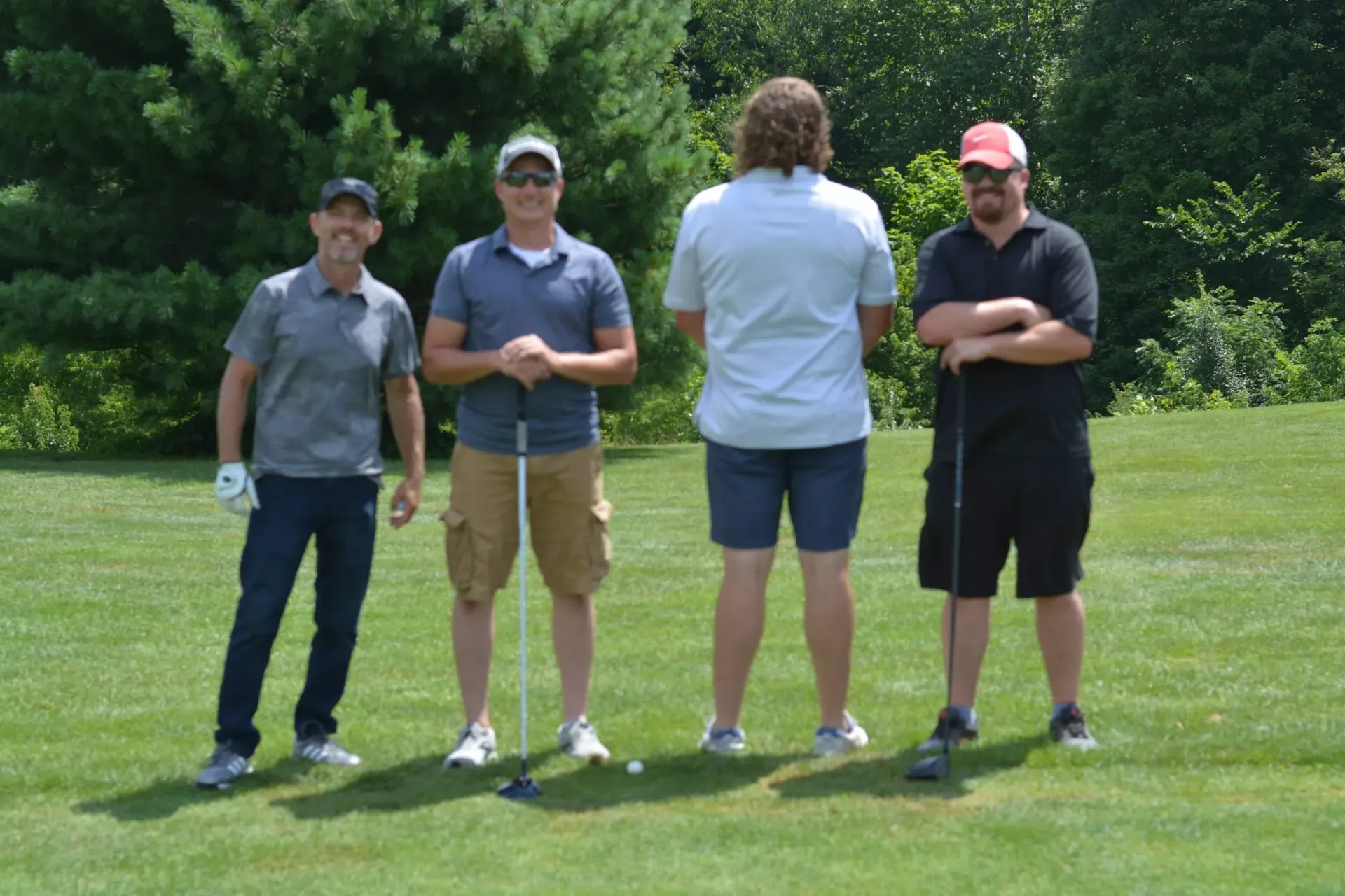Four people standing on a grassy field, three facing the camera and one turned away, with trees in the background.