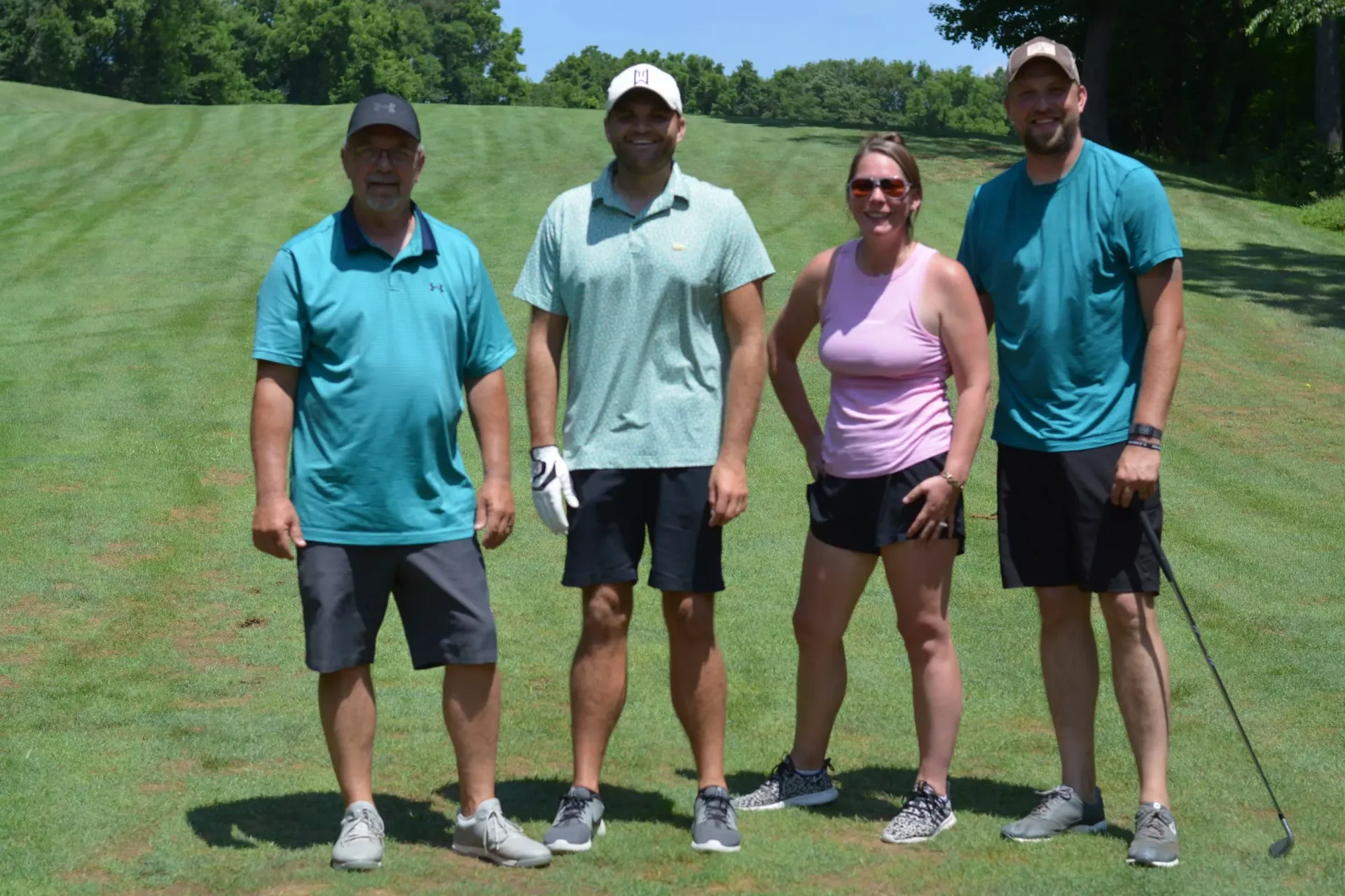 Four golfers stand together on a grassy course, holding clubs and posing for a photo.