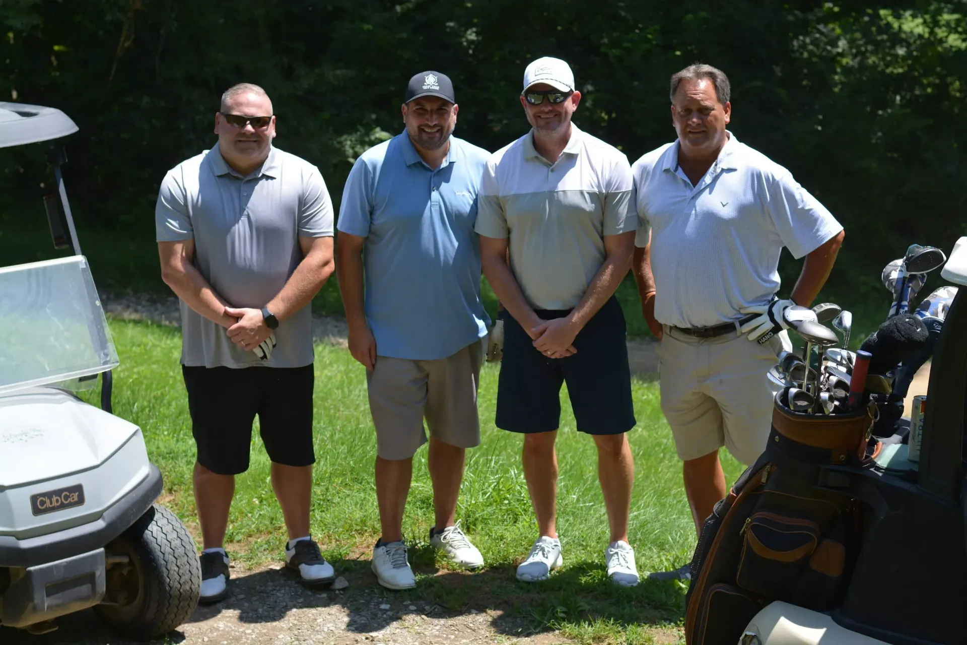 Four golfers standing on grass beside golf carts, posing outdoors in bright sunlight.