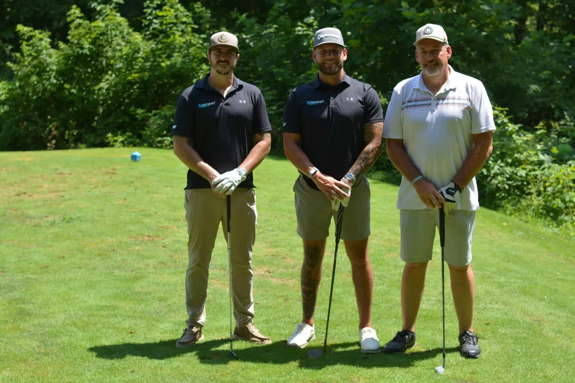 Three golfers standing on a sunny green course, holding clubs and posing side by side.