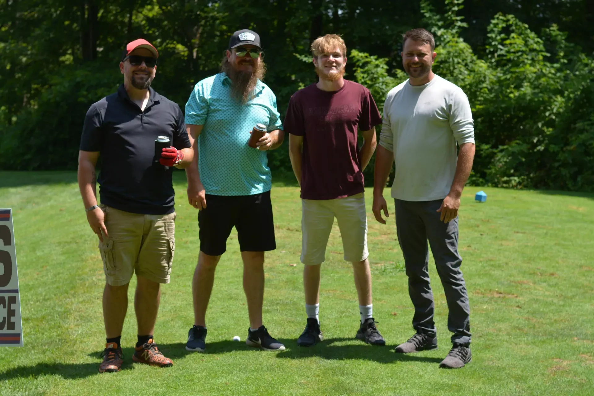 Four men standing on a sunny golf course lawn, holding drinks and smiling.