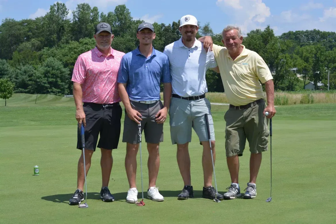 Four golfers standing together on a green golf course, holding clubs, with trees in the background.