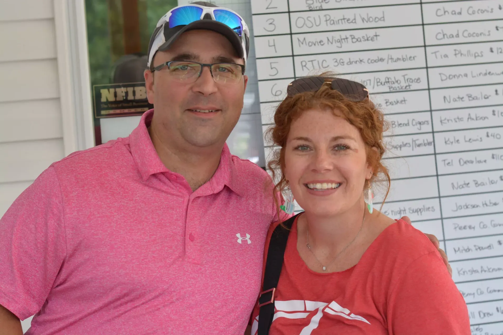 Two smiling people standing together outdoors, wearing pink shirts, with a signboard behind them