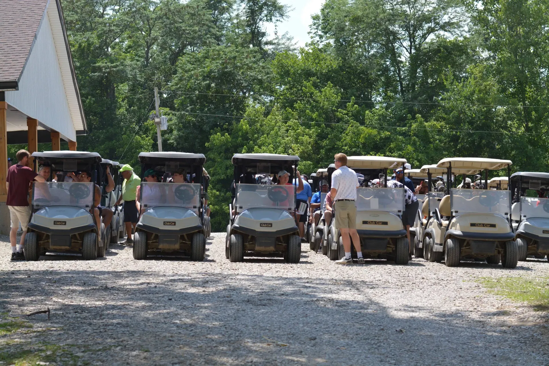 Row of golf carts parked on a gravel path under trees, with people standing beside them