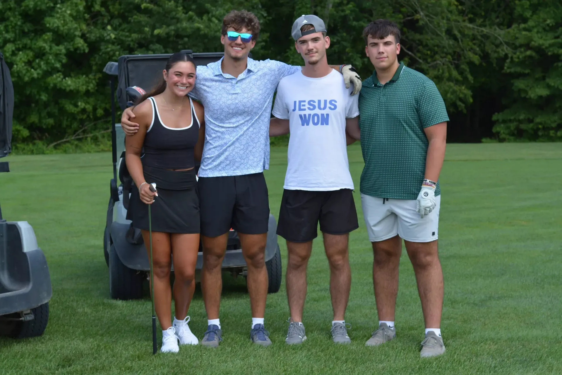 Four friends standing on grass, smiling arm-in-arm, with a golf cart in the background