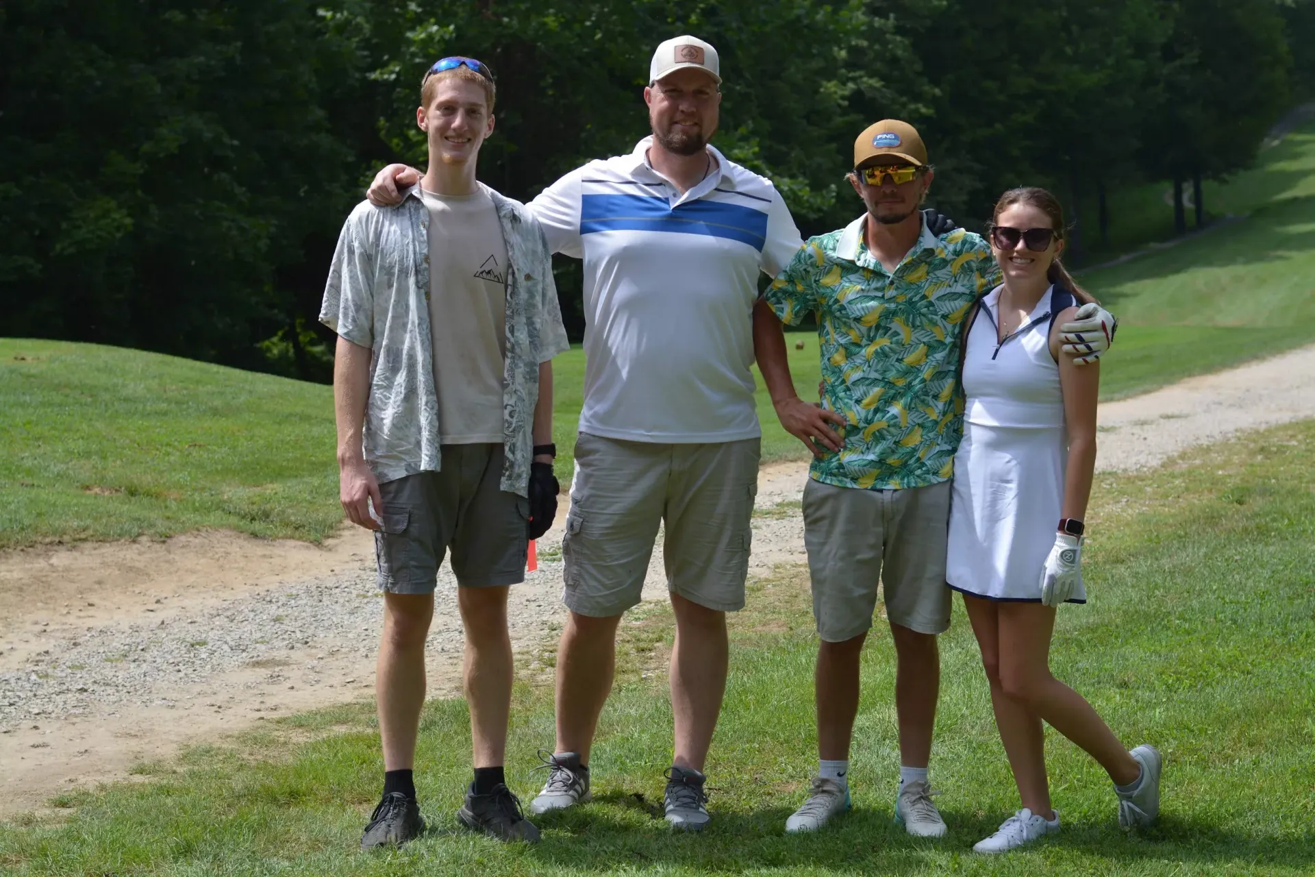 Four people standing on grass by a golf course path, posing together outdoors.