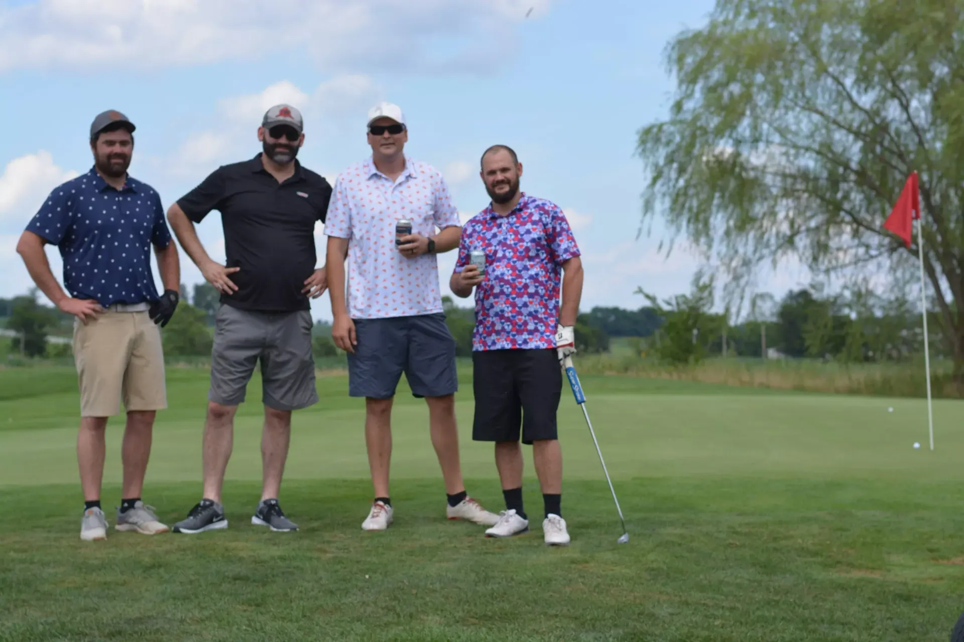 Four golfers posing on a green beside a golf flag under a cloudy sky.
