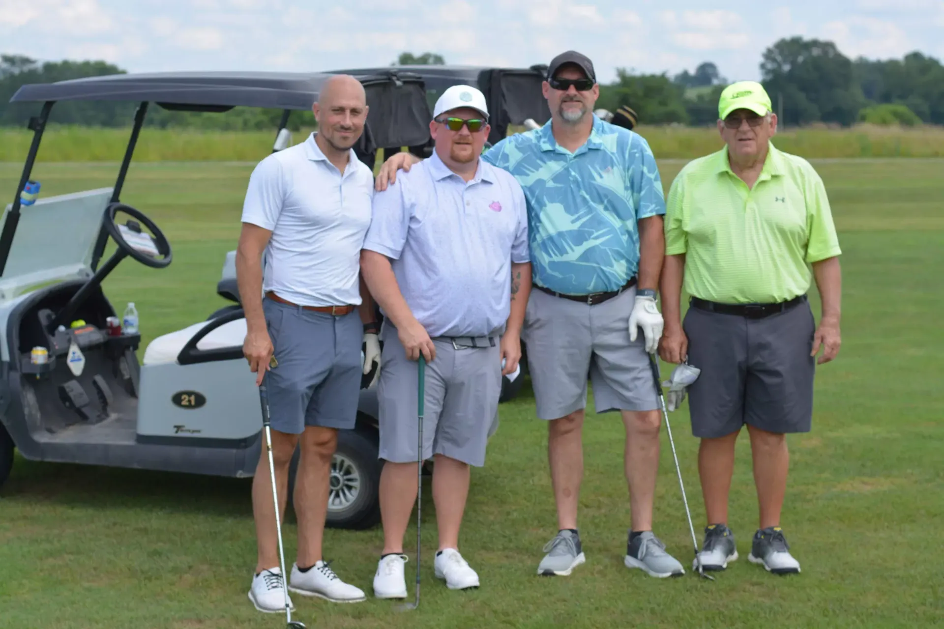 Four men standing beside a golf cart on a grassy golf course, posing for a photo.