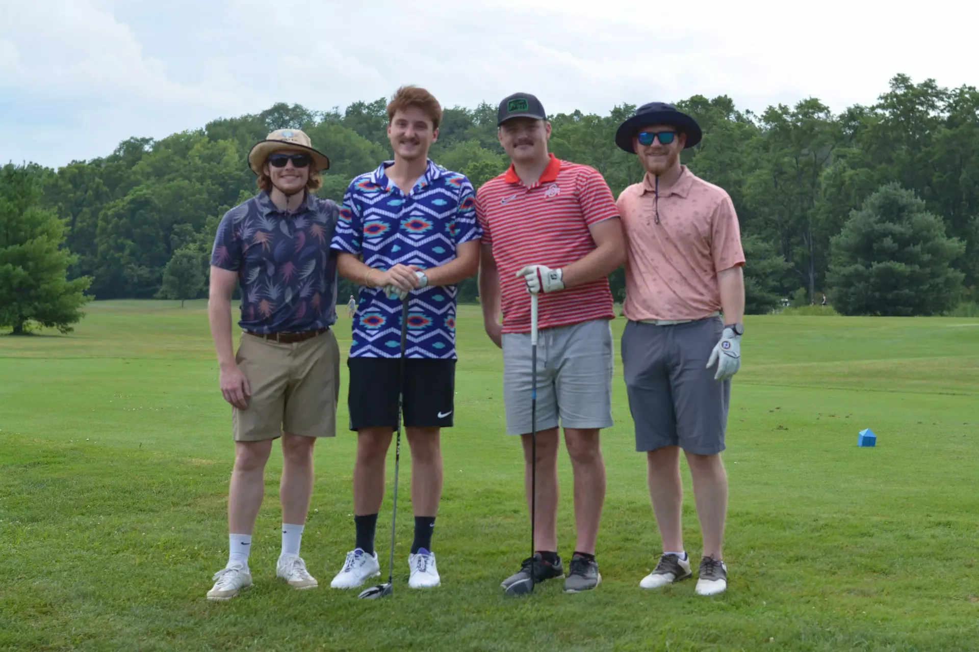Four people posing with golf clubs on a grassy course, with trees in the background.