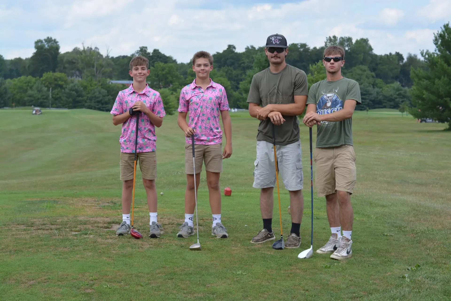 Four golfers standing on a grassy course, holding clubs and posing before a shot.