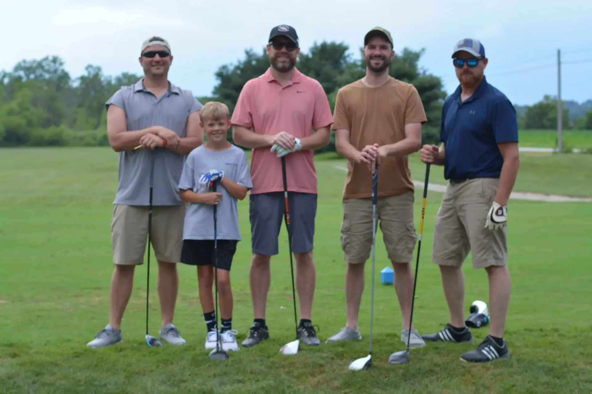Five people posing on a golf course, holding clubs and smiling at the camera.