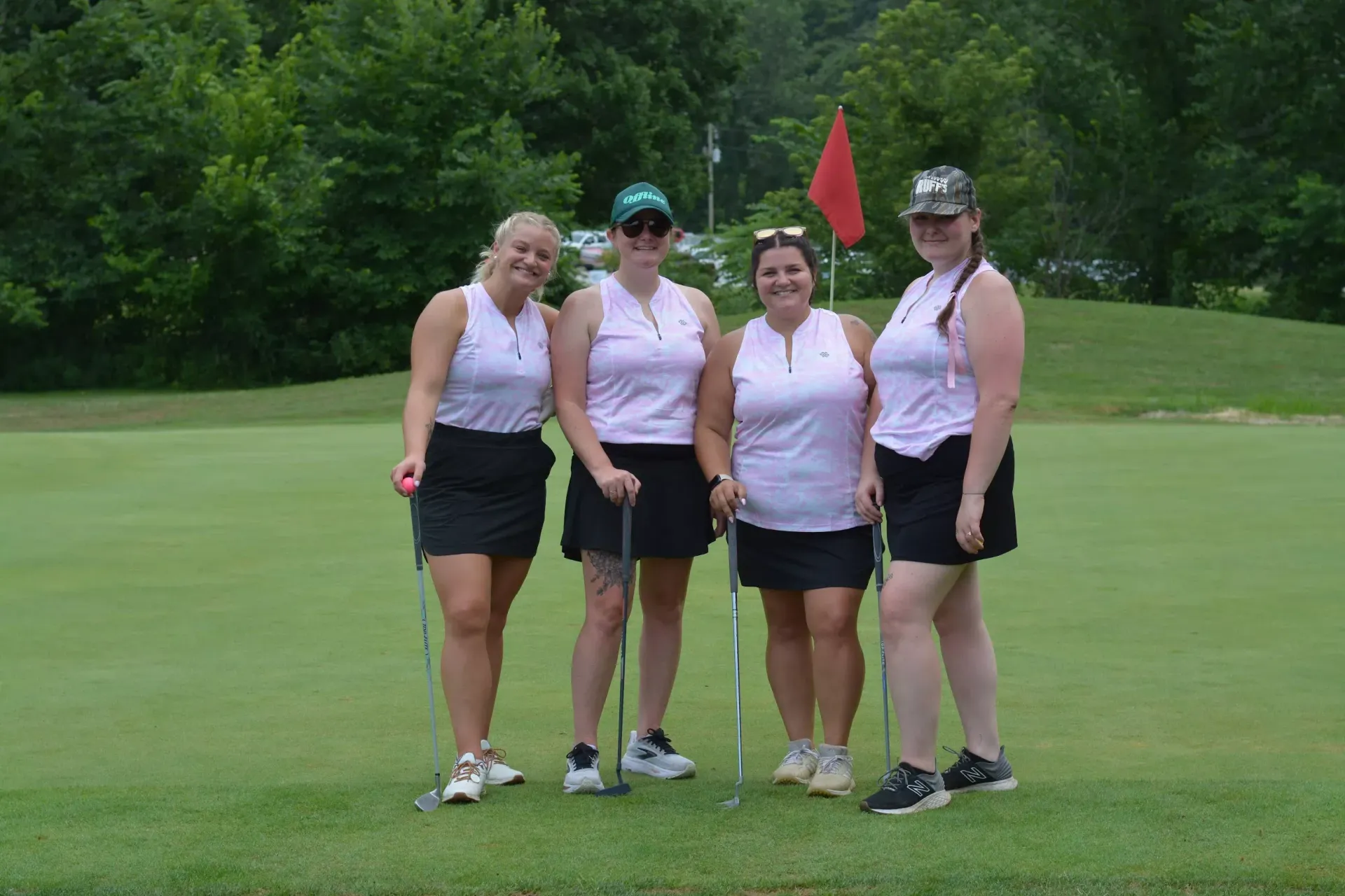 Four golfers in white tops and dark skirts posing on a green golf course near a red flag