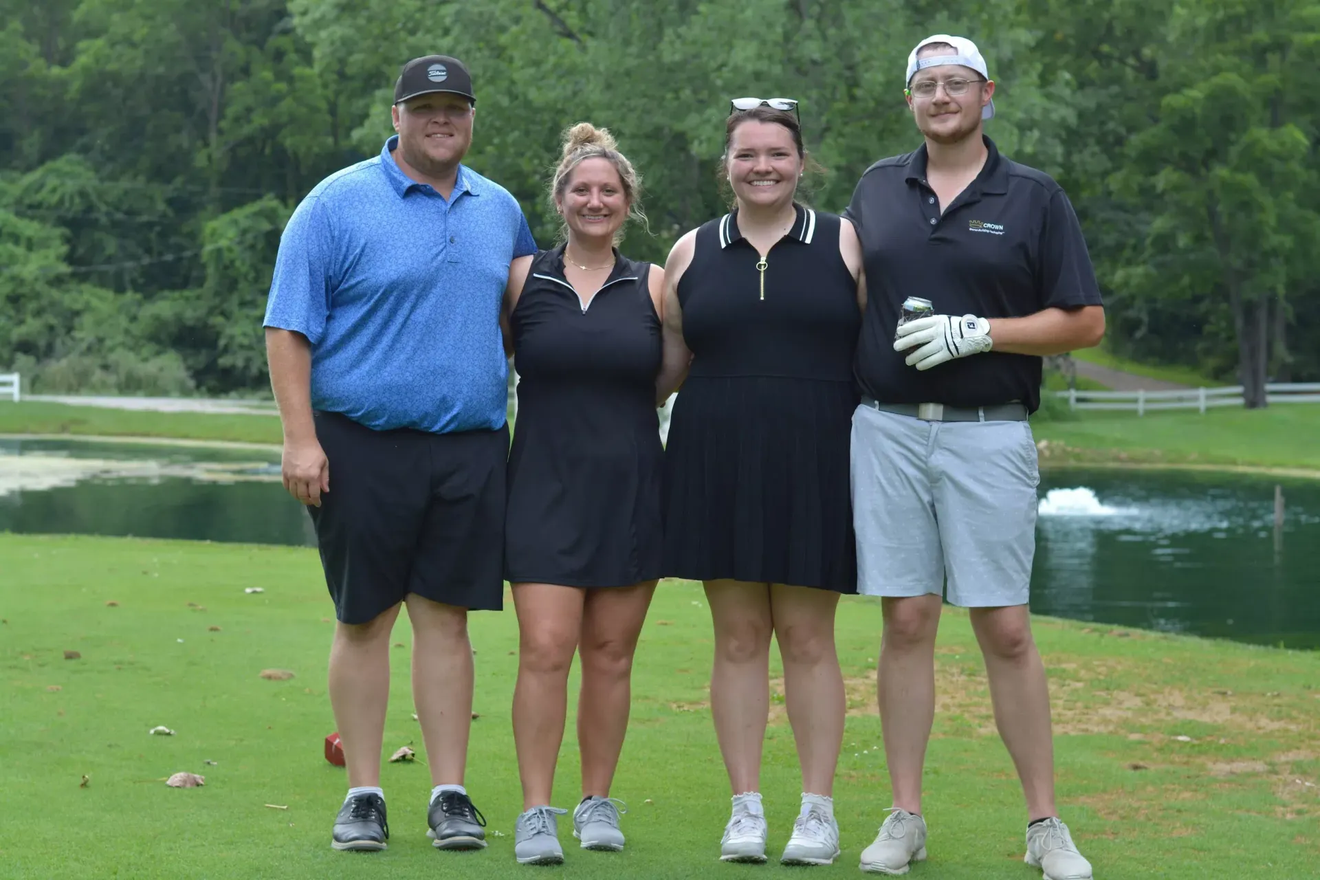 Four golfers posing on a green beside a pond, with trees in the background.