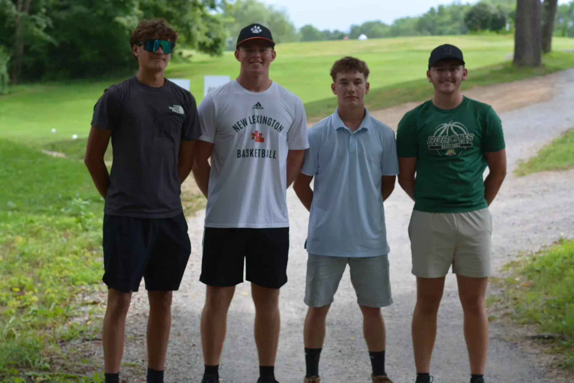 Four friends stand on a gravel path in a park, with trees and green fields in the background.