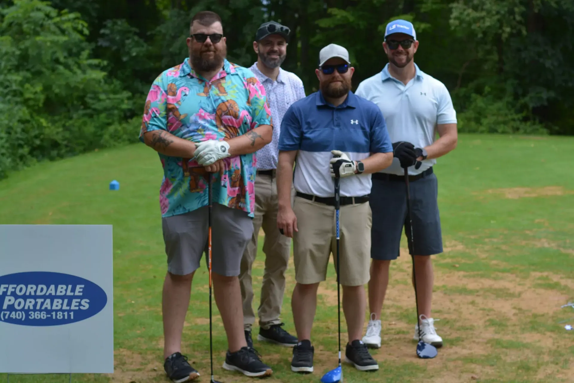 Four men posing on a golf course beside a sign, holding clubs and wearing casual golf attire