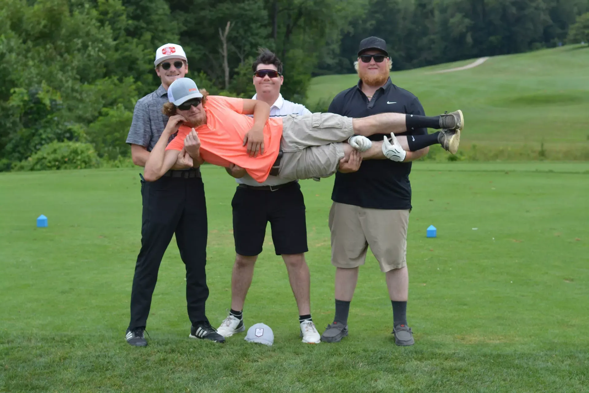 Three people carrying a man horizontally on a golf course grassy fairway