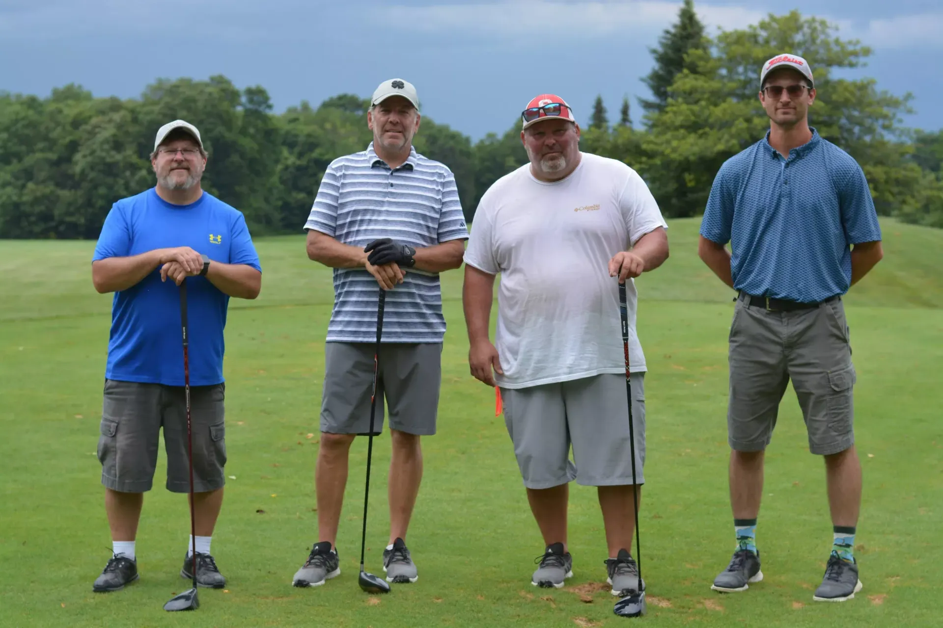 Four golfers standing on a green tee box, holding clubs with trees and cloudy sky behind them