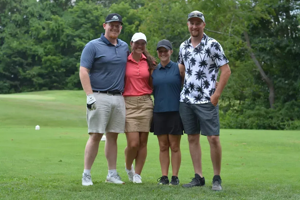 Four people posing together on a golf course, standing on the green with trees in the background.