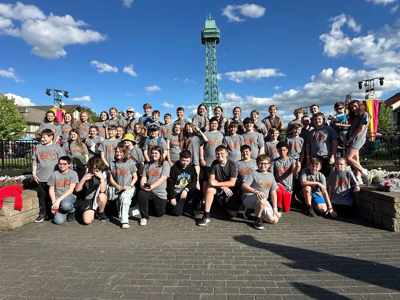 Large group posing outdoors in front of a tall tower under a blue sky, with colorful flags on the right.