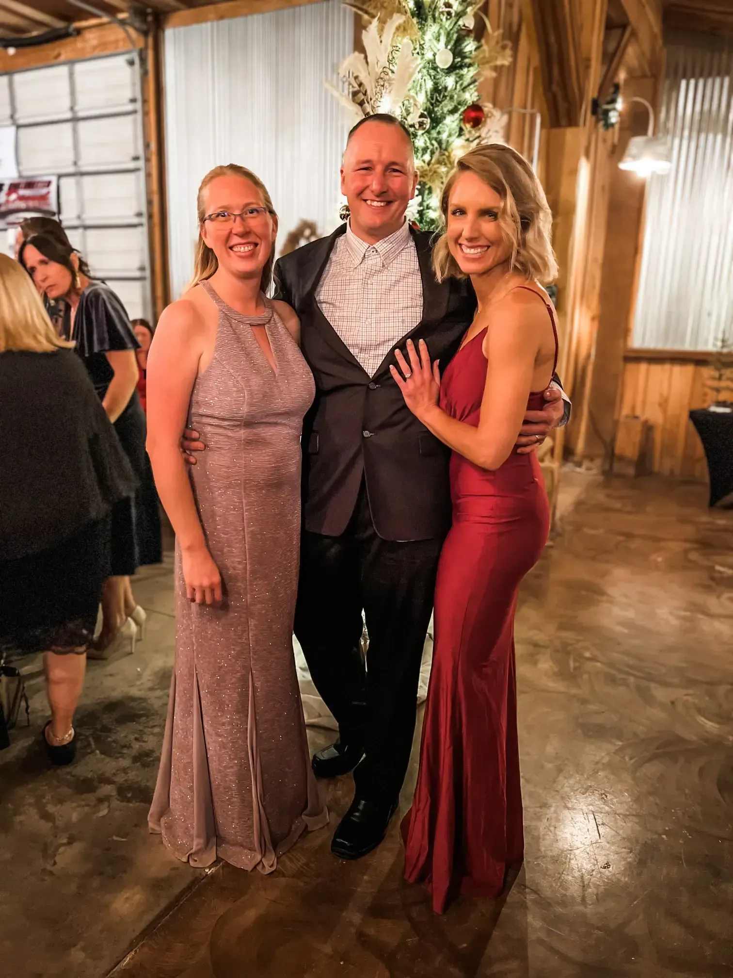 Three people in formal attire smiling at a rustic indoor event.