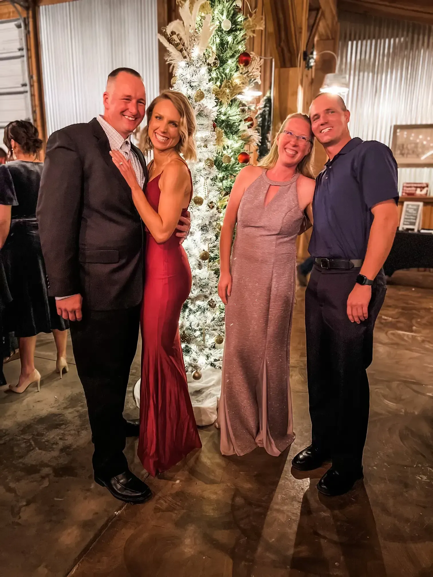 Four people posing and smiling beside a lit Christmas tree at an indoor party.