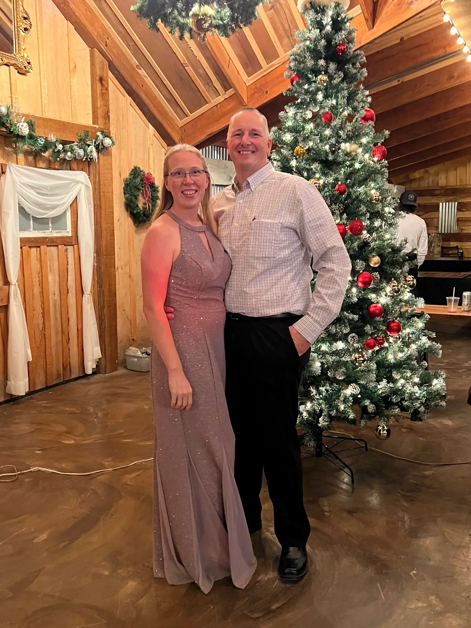 Couple posing in a wood-paneled hall beside a decorated Christmas tree.