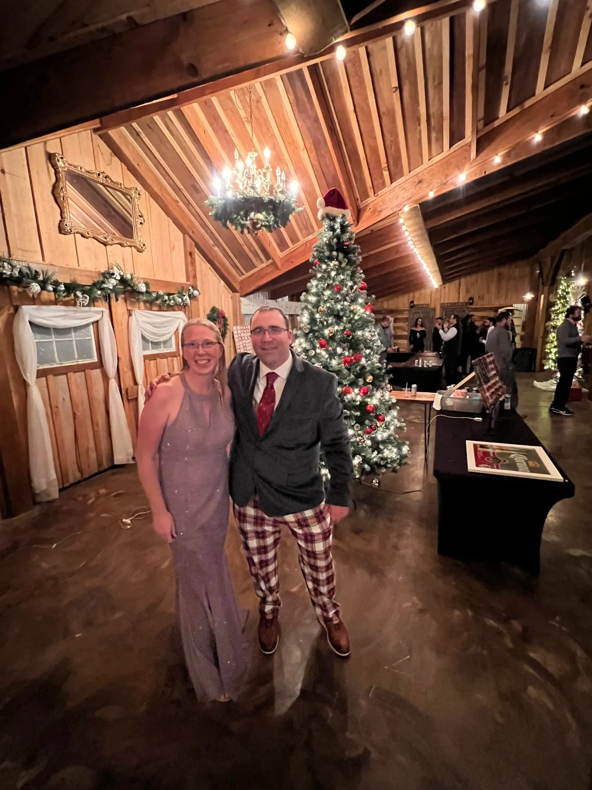 Two people posing in a festive hall beside a decorated Christmas tree, one in a pink gown and one in a suit