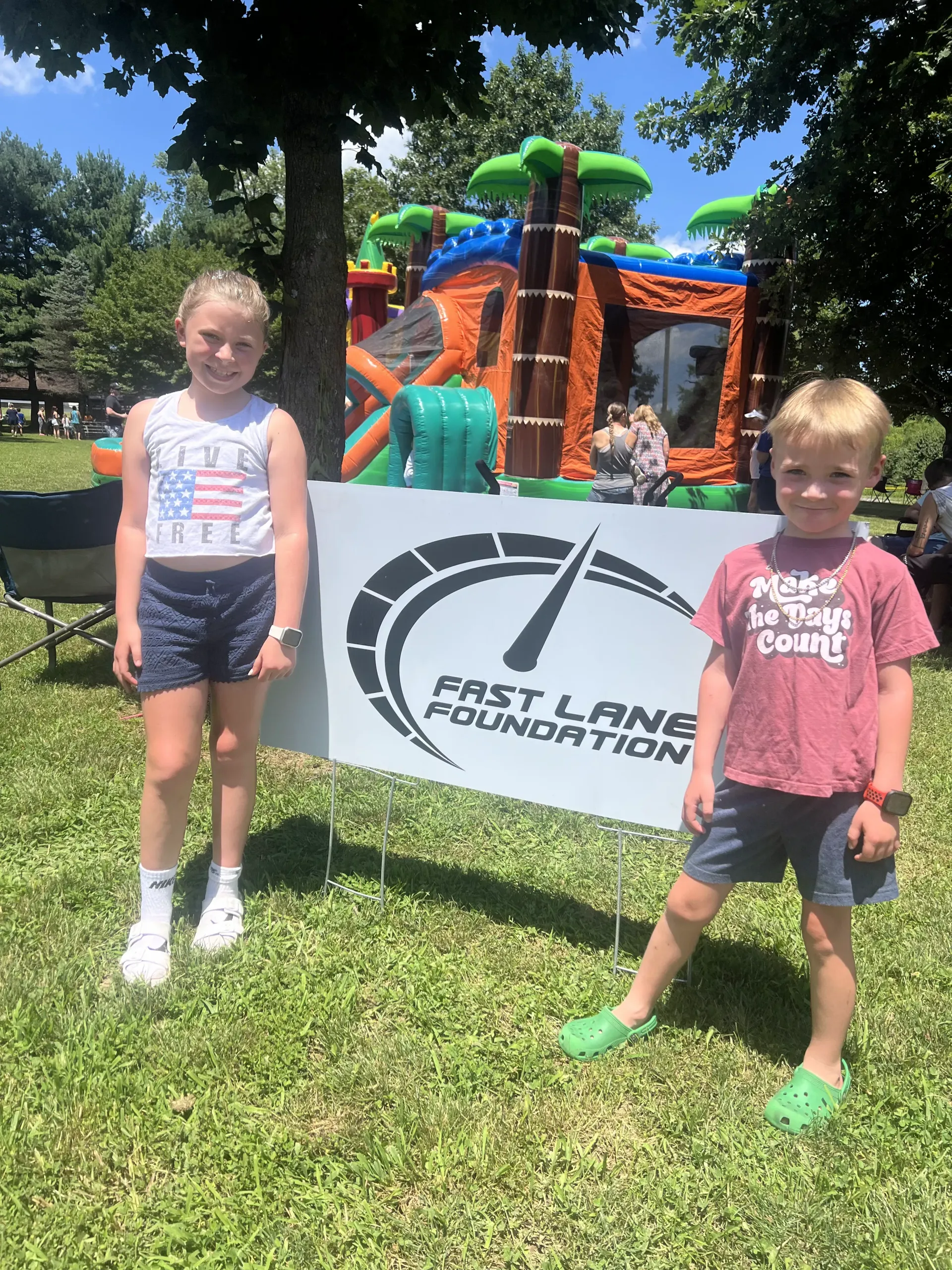 Two children standing on grass in front of a bouncy castle and a First Lane Foundation sign