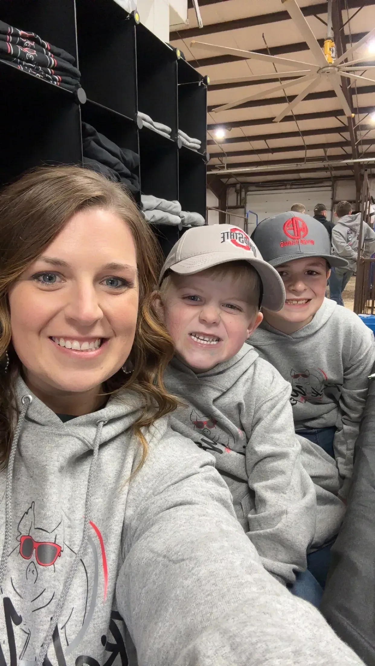 Three people in gray hoodies smiling in a warehouse aisle, taking a selfie among stacked black bins.