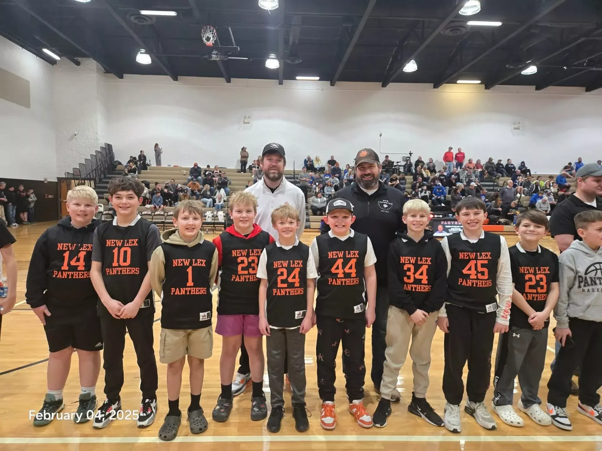 Youth basketball team posing in black and orange jerseys with coaches in a gym, crowd seated in bleachers behind them