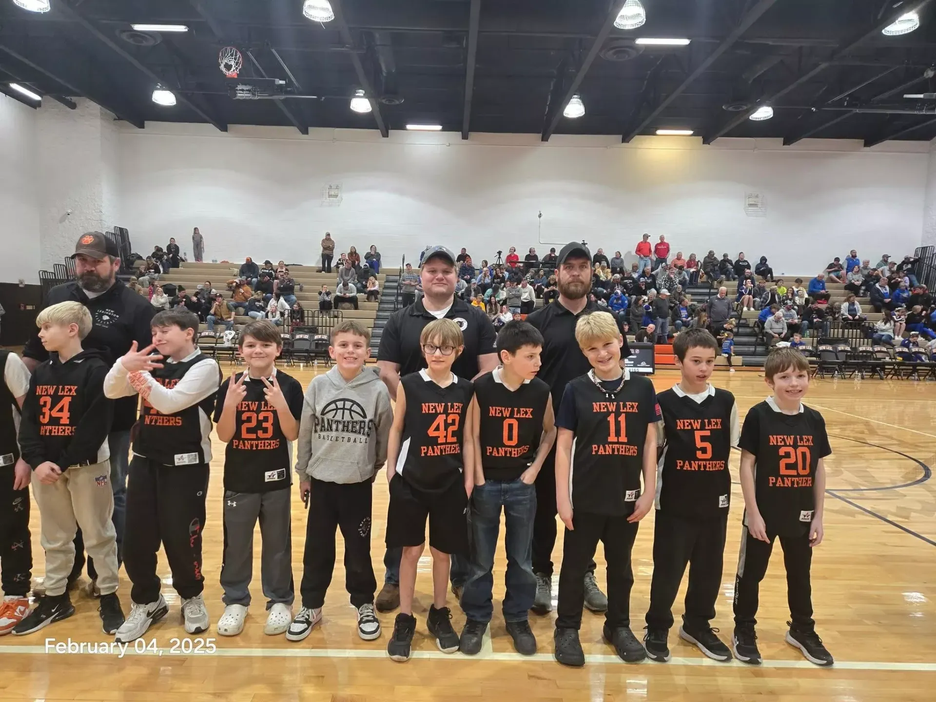 Youth basketball team lined up on court with coaches and crowd in gymnasium, wearing black jerseys with orange numbers