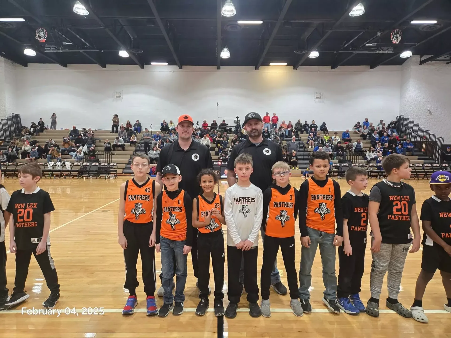 Youth basketball team posing on court in orange and black uniforms with coaches in a gymnasium