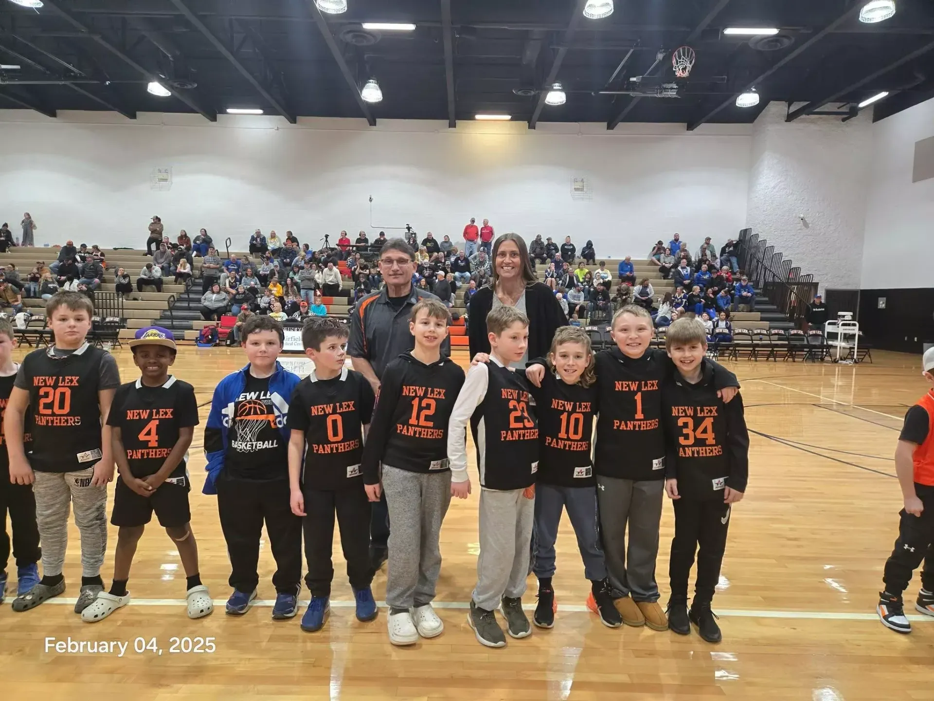 Youth basketball team posing on a gym court during a game, wearing black and orange jerseys.