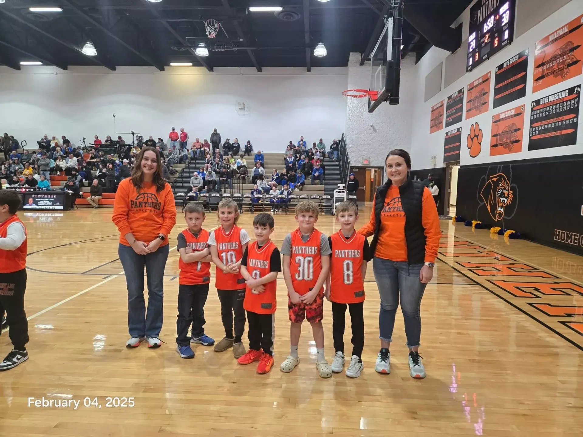 Youth basketball team in orange jerseys and coaches posing on a gym court during a game.