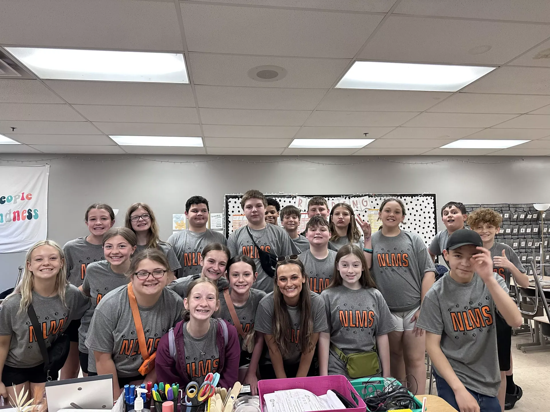 Group of students in gray shirts posing in a classroom, smiling behind a table of colorful items.