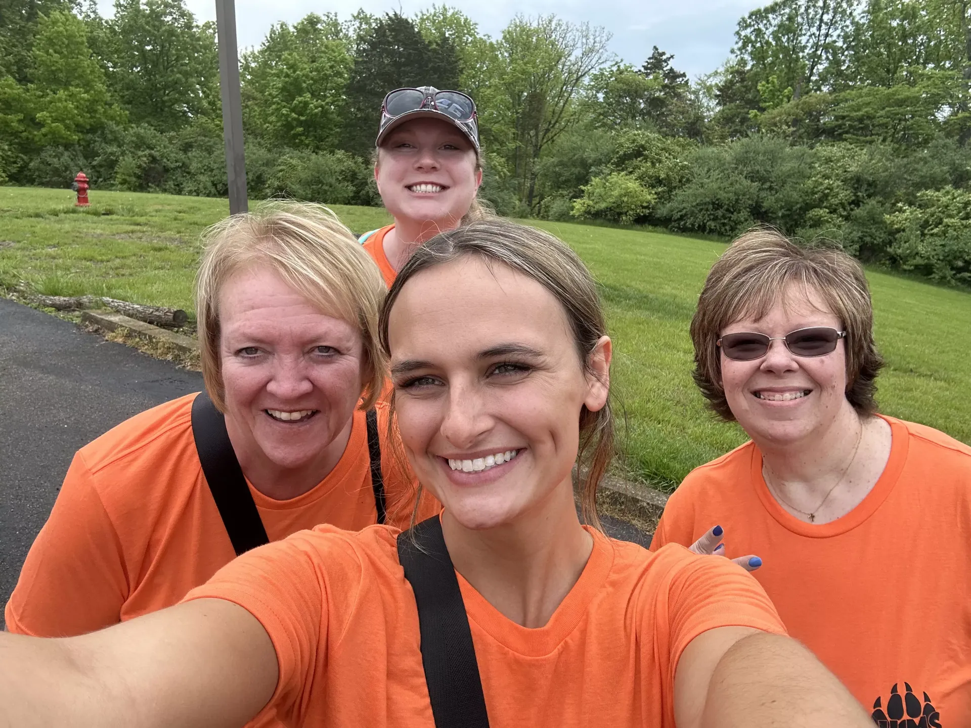 Four people in orange shirts smile outdoors on a grassy trail with trees in the background.